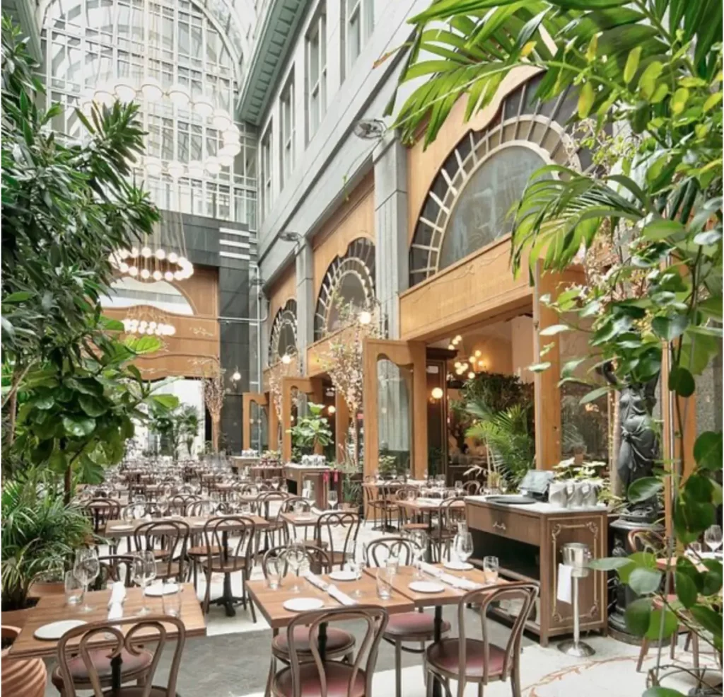 Intérieur d'un restaurant new-yorkais spacieux, baigné de lumière naturelle grâce à une verrière imposante, avec une décoration luxuriante mêlant plantes vertes et mobilier en bois élégant