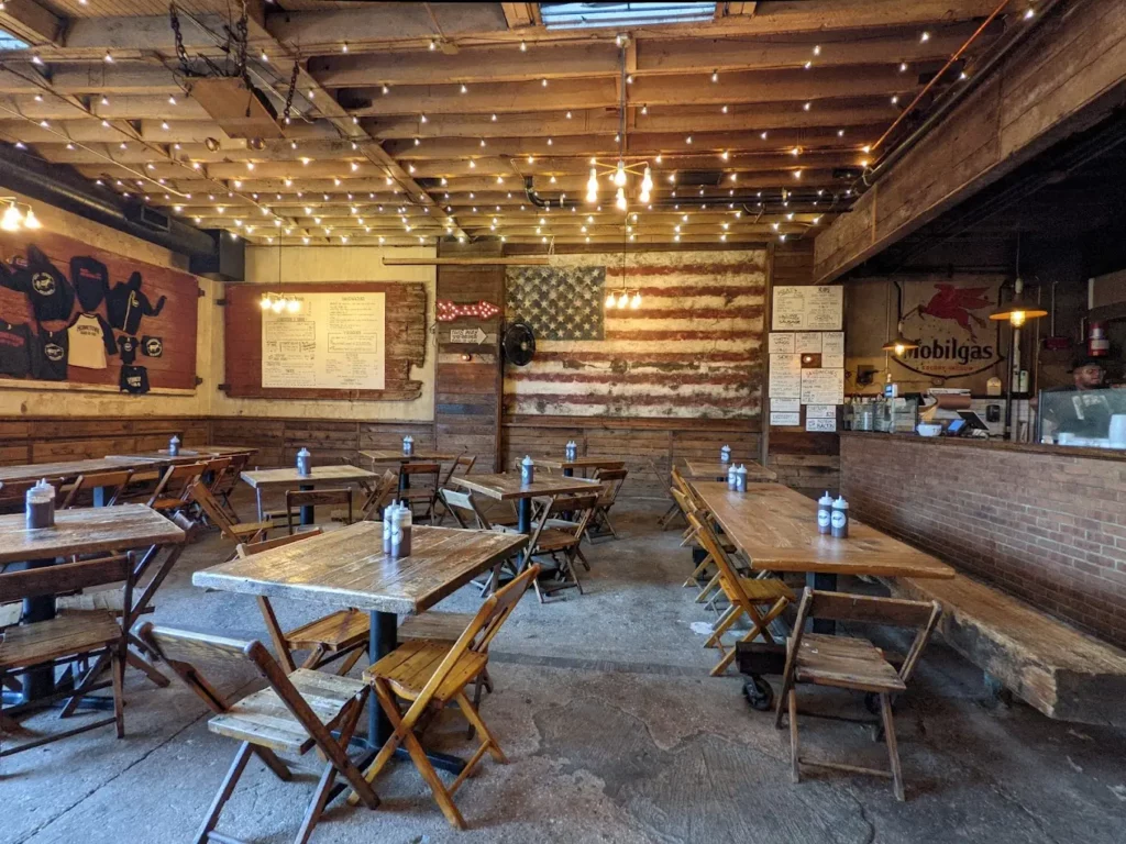 Intérieur d'un restaurant new-yorkais au style rustique, avec des tables en bois, des chaises pliantes et une décoration vintage mettant en avant un drapeau américain mural.