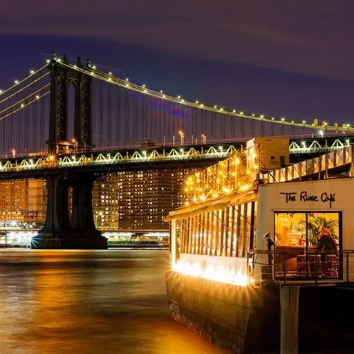 Restaurant new-yorkais en bord de rivière, illuminé en soirée, avec une vue imprenable sur un pont emblématique de la ville.